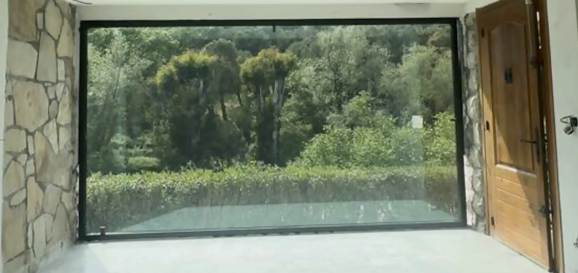 Vue depuis l’intérieur à travers une grande fenêtre rectangulaire vers des arbres et des arbustes.