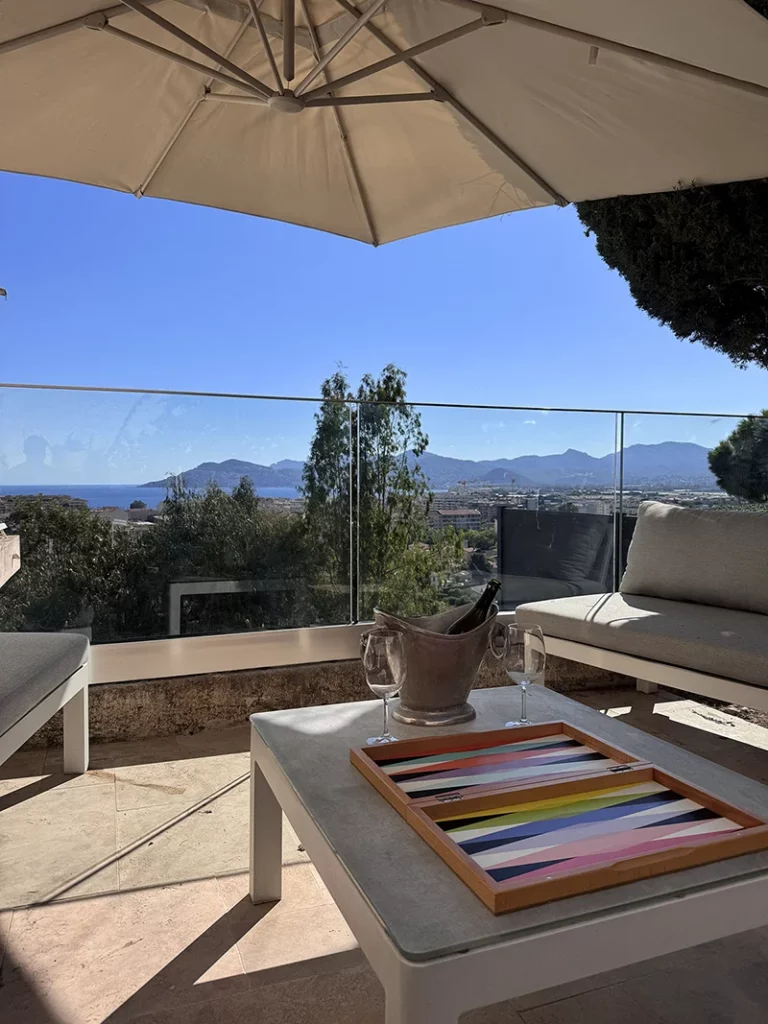 Terrasse ensoleillée avec garde-corps transparent, mobilier extérieur et vue sur l’eau et les montagnes.
