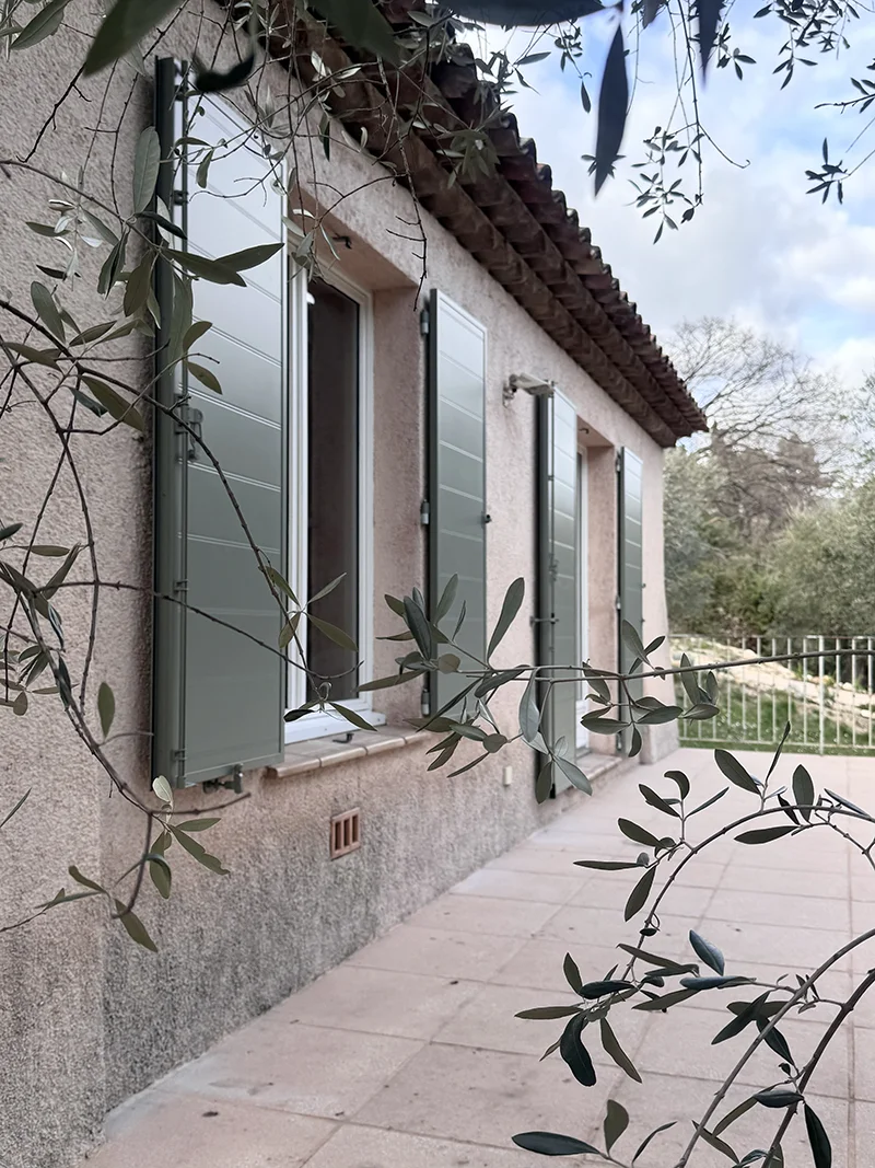 Façade en stuc avec plusieurs fenêtres aux volets gris-vert ouverts, branches au premier plan et terrasse pavée.