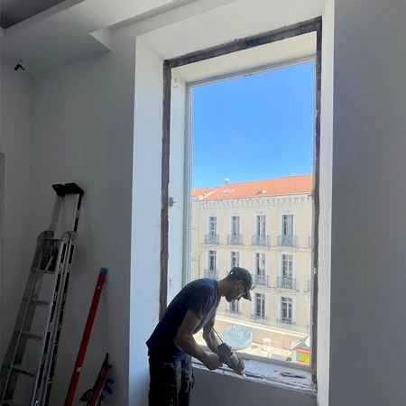 Une personne travaille près d’une fenêtre en rénovation, avec une échelle et des outils contre le mur.