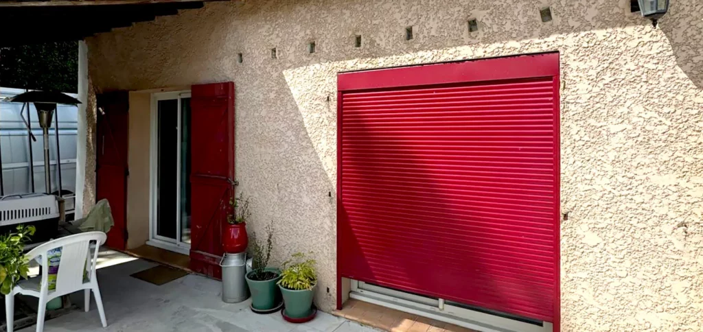 Façade beige avec grand volet roulant rouge fermé, porte rouge et plantes en pots.