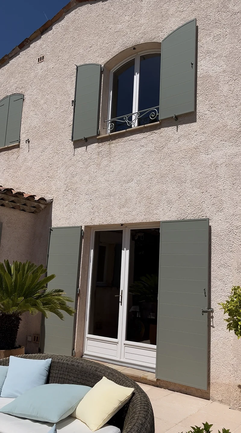Façade en stuc avec volets, portes vitrées et espace patio aménagé.