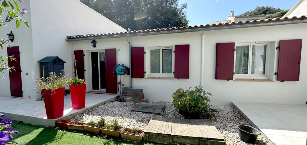 Maison blanche de plain-pied avec volets et portes bordeaux autour d’une cour paysagée