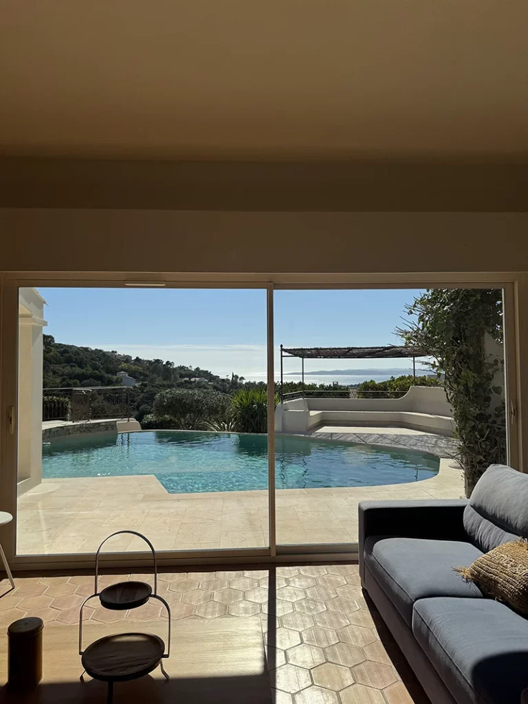 Intérieur lumineux ouvrant sur une piscine, une terrasse et un paysage vallonné par de grandes baies vitrées coulissantes.